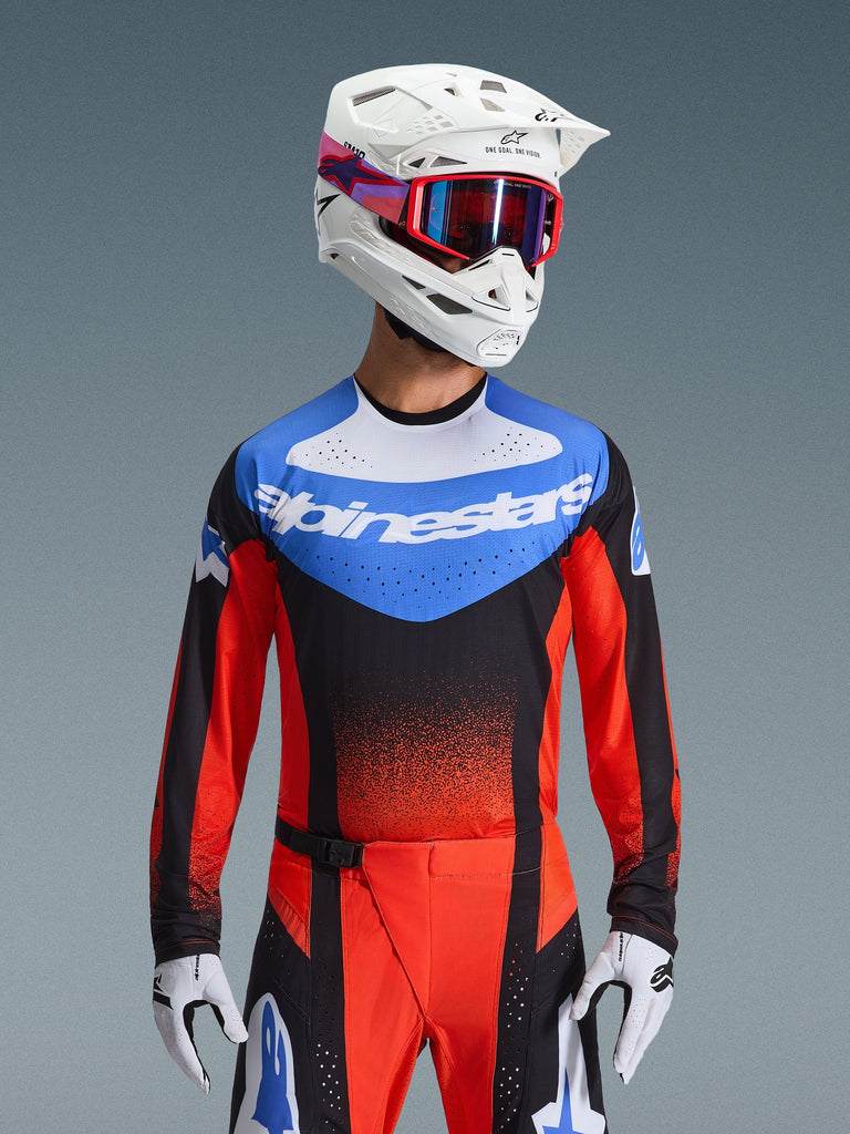 Alpinestars Techstar Knif Jersey 2026, maillot de motocross, orange, noir et bleu UCLA, avec empiècement bleu sur la poitrine, logo Alpinestars blanc et manches orange-rouge, porté avec un casque blanc, des lunettes rouges et des gants blancs, équipement de course tout-terrain professionnel