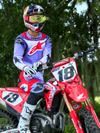 Pilote de motocross sur une moto Honda CRF rouge, portant le maillot Alpinestars 2025 Fluid Wurx violet et rouge vif, pantalon assorti violet et rouge, casque Red Bull avec lunettes jaunes, bottes et gants Alpinestars blancs, équipement de motocross professionnel pour le tout-terrain