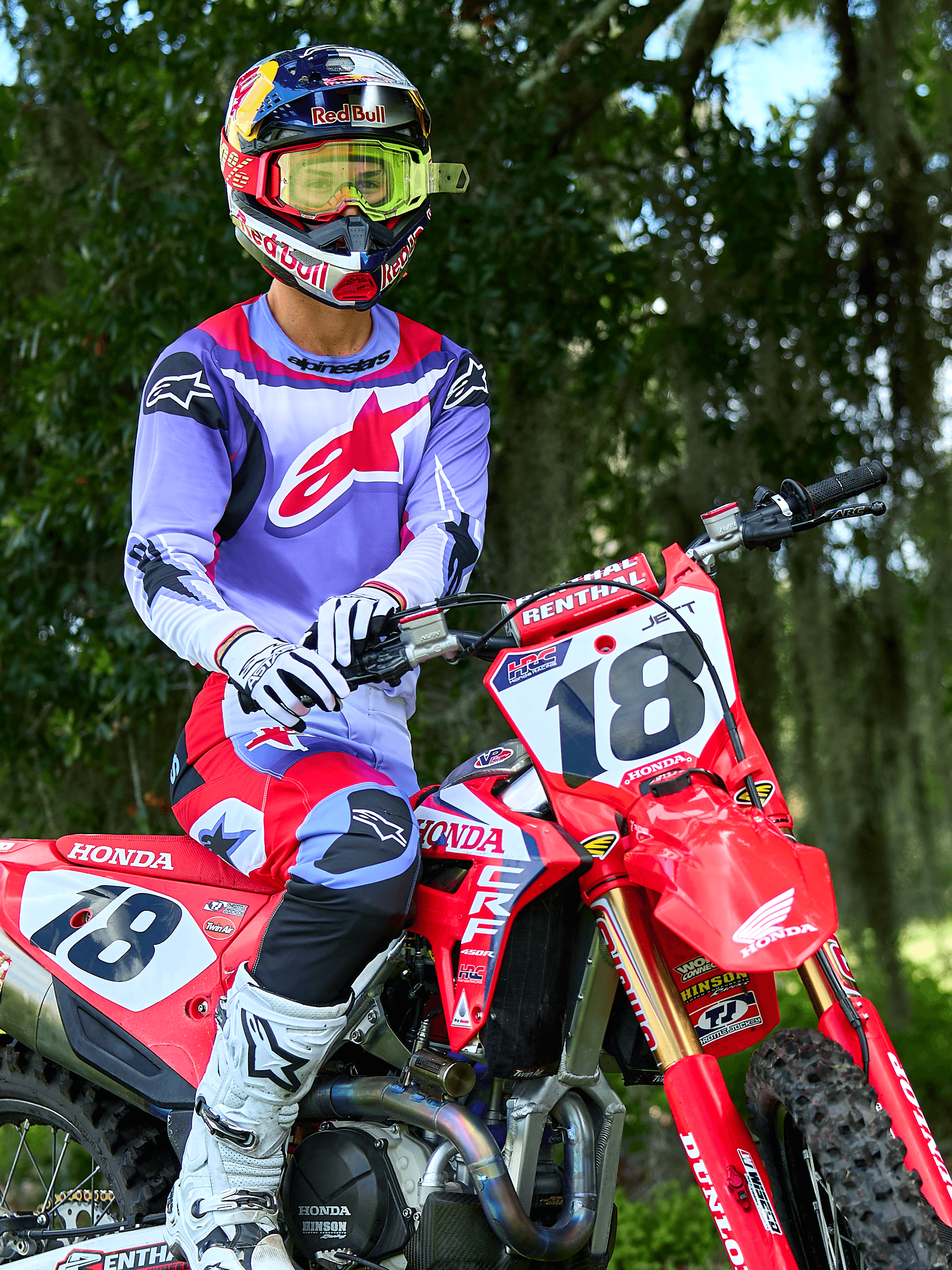 Pilote de motocross sur une moto Honda CRF rouge, portant le maillot Alpinestars 2025 Fluid Wurx violet et rouge vif, pantalon assorti violet et rouge, casque Red Bull avec lunettes jaunes, bottes et gants Alpinestars blancs, équipement de motocross professionnel pour le tout-terrain