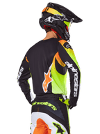 Motocross rider from behind wearing the Alpinestars 2025 Fluid Wurx Gear Set, black jersey with vibrant neon yellow and orange graphics, matching pants with white and orange panels, black helmet with neon accents, bold Alpinestars branding on sleeves and seat