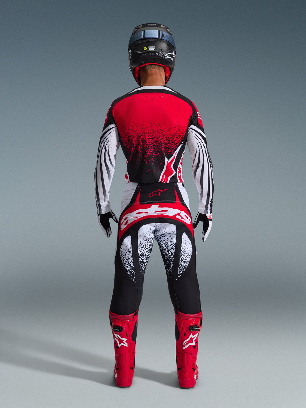 Motocross rider from behind wearing the Alpinestars 2026 Techstar Nomur Gear Set, red and white jersey with black speckle gradient, matching white and red pants with black accents, bright red boots, and black helmet, coordinated off-road racing gear.