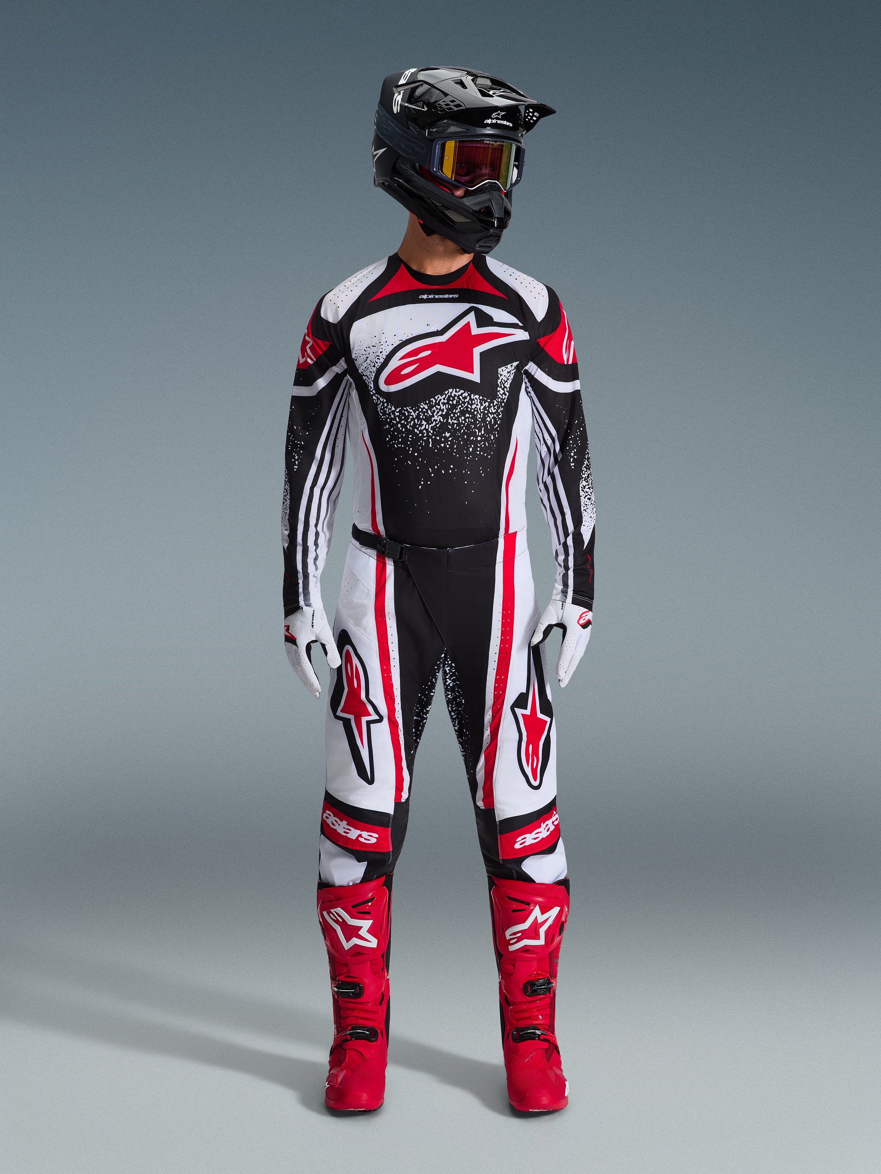 Pilote de motocross portant la tenue Techstar Nomur 2026, noir, blanc et rouge, maillot arborant un grand logo étoile rouge et un motif moucheté, pantalon assorti avec des panneaux latéraux rouges, casque noir avec lunettes teintées et bottes de motocross rouges.