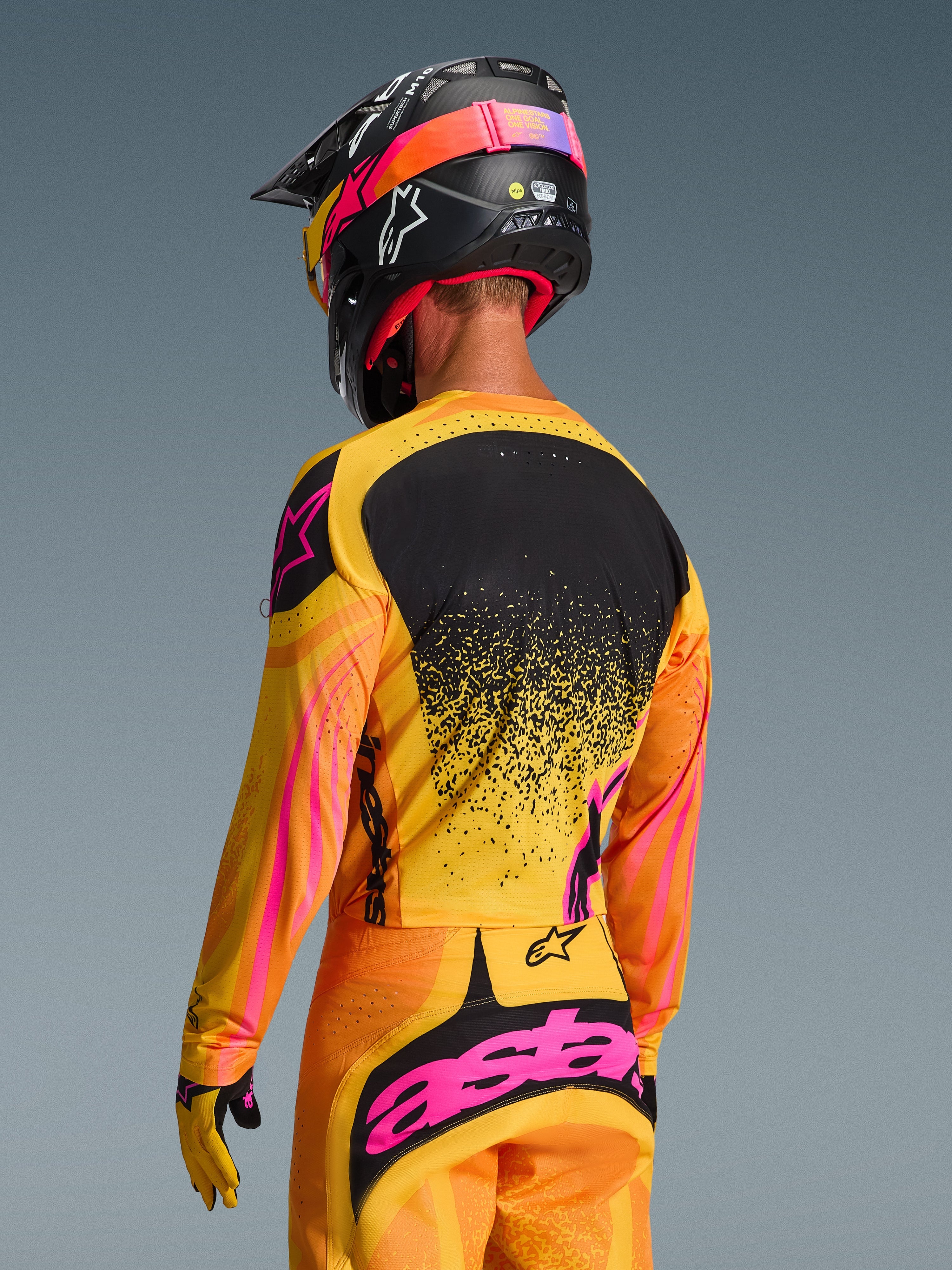 Maillot Alpinestars Techstar Nomur 2026 en Orange Jaune Rose, vue arrière d'un pilote de motocross portant une tenue complète avec pantalon assorti et casque noir, design dynamique aux accents jaunes et roses et panneau arrière noir, coupe athlétique pour la performance tout-terrain