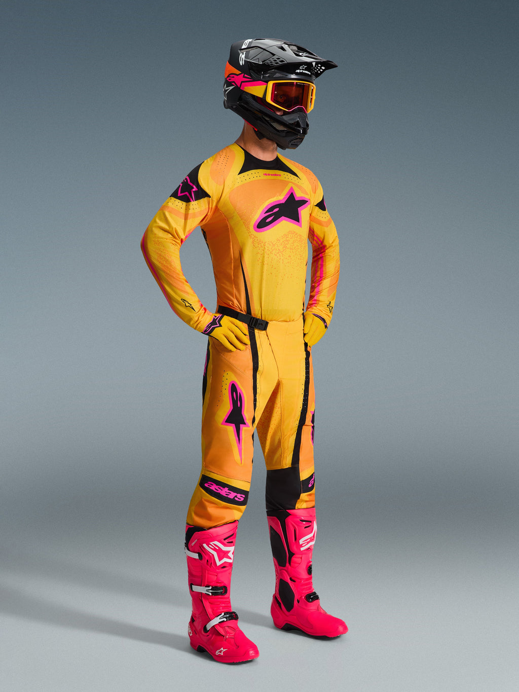 Pilote de motocross portant le maillot Techstar Nomur 2026 en orange, jaune et rose, pantalon orange assorti avec des accents roses, bottes rose vif, gants jaunes et casque noir avec lunettes jaunes, tenue de motocross coordonnée
