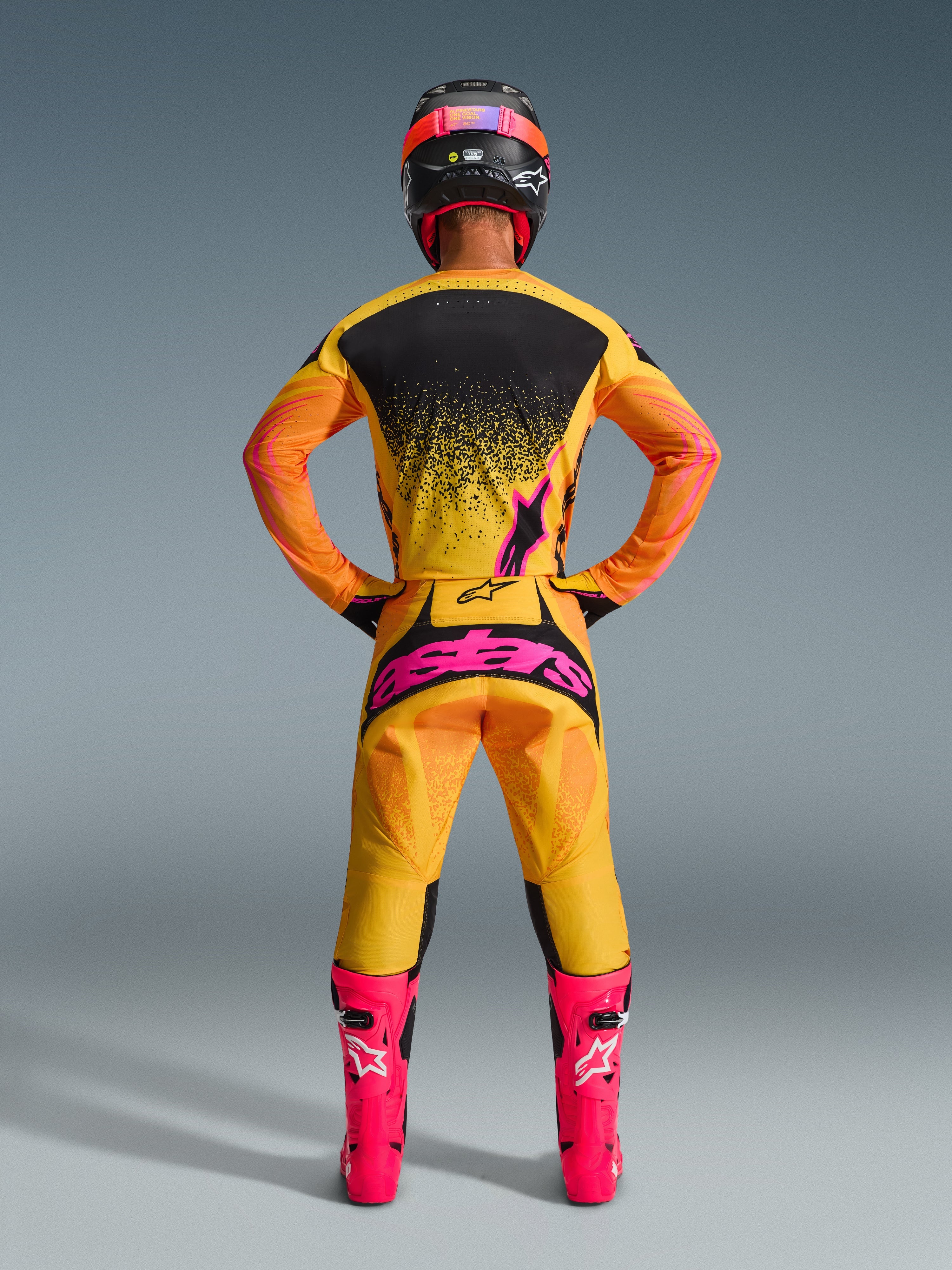 Pilote de motocross de dos portant le maillot Alpinestars 2026 Techstar Nomur en Orange Jaune Rose, tenue de course coordonnée avec pantalon assorti au logo rose vif et bottes rose fluo, coupe athlétique pour performance tout-terrain de niveau avancé.