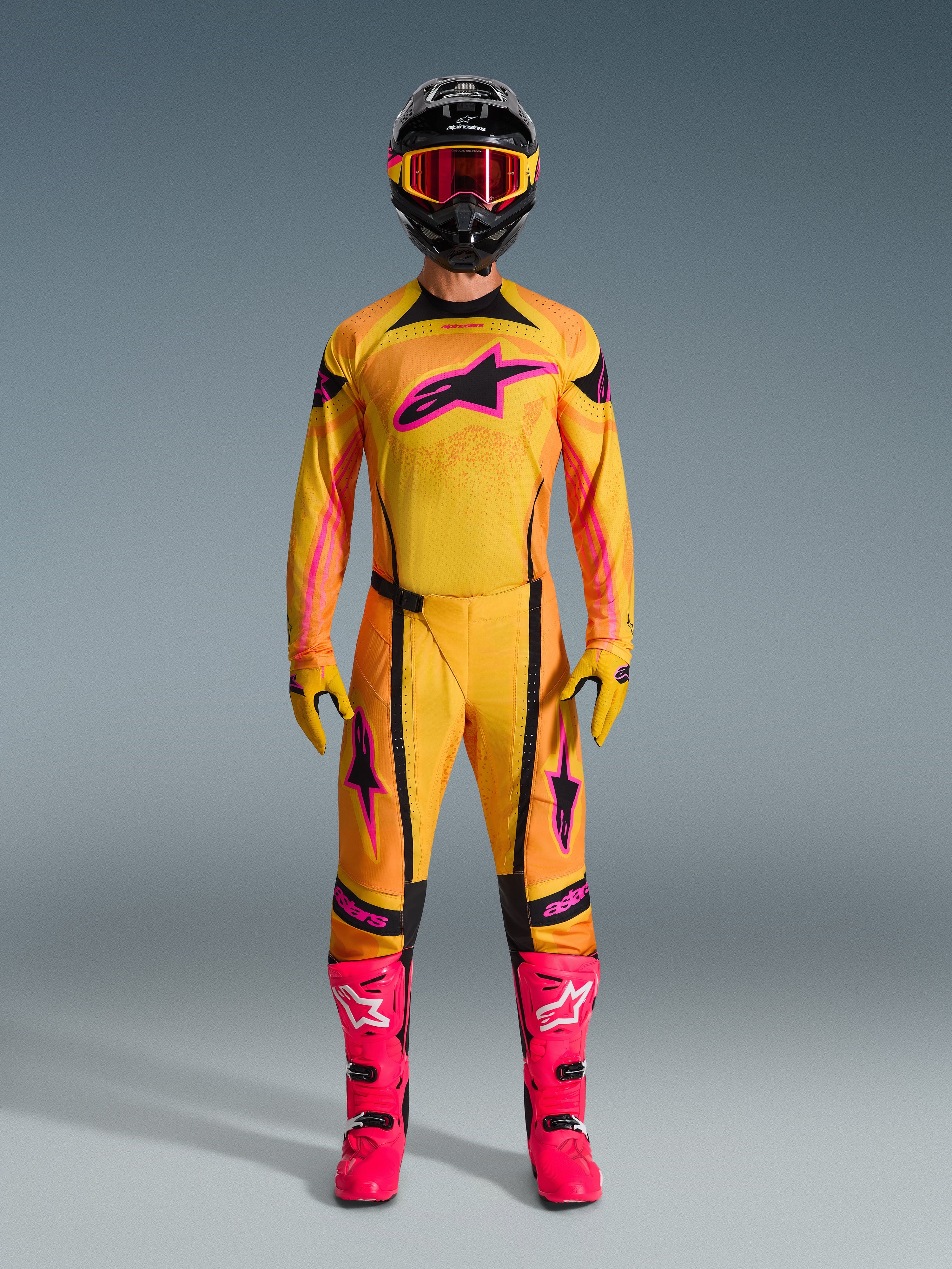 Pilote de motocross portant le maillot Techstar Nomur 2026 en orange, jaune et rose, coordonné avec un pantalon de motocross assorti, un casque noir avec des lunettes roses et des bottes rose vif, équipement de course athlétique pour la compétition tout-terrain