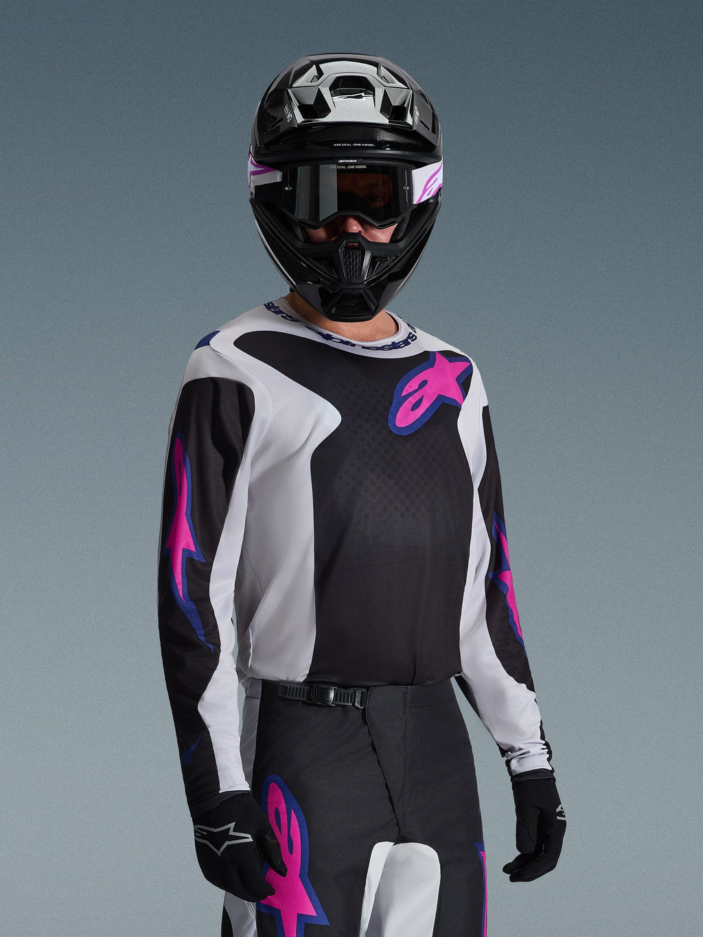 Pilote de motocross portant le maillot Alpinestars Fluid Grid 2026, noir, gris clair et violet, avec de grands logos étoilés roses et violets, accompagné d'un pantalon assorti, d'un casque noir et de lunettes, tenue de course tout-terrain complète