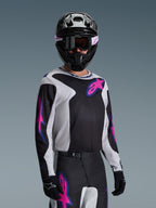 Pilote de motocross portant le maillot Alpinestars Fluid Grid 2026, noir, gris clair et violet, avec de grands logos étoilés roses et violets, accompagné d'un pantalon assorti, d'un casque noir et de lunettes, tenue de course tout-terrain complète