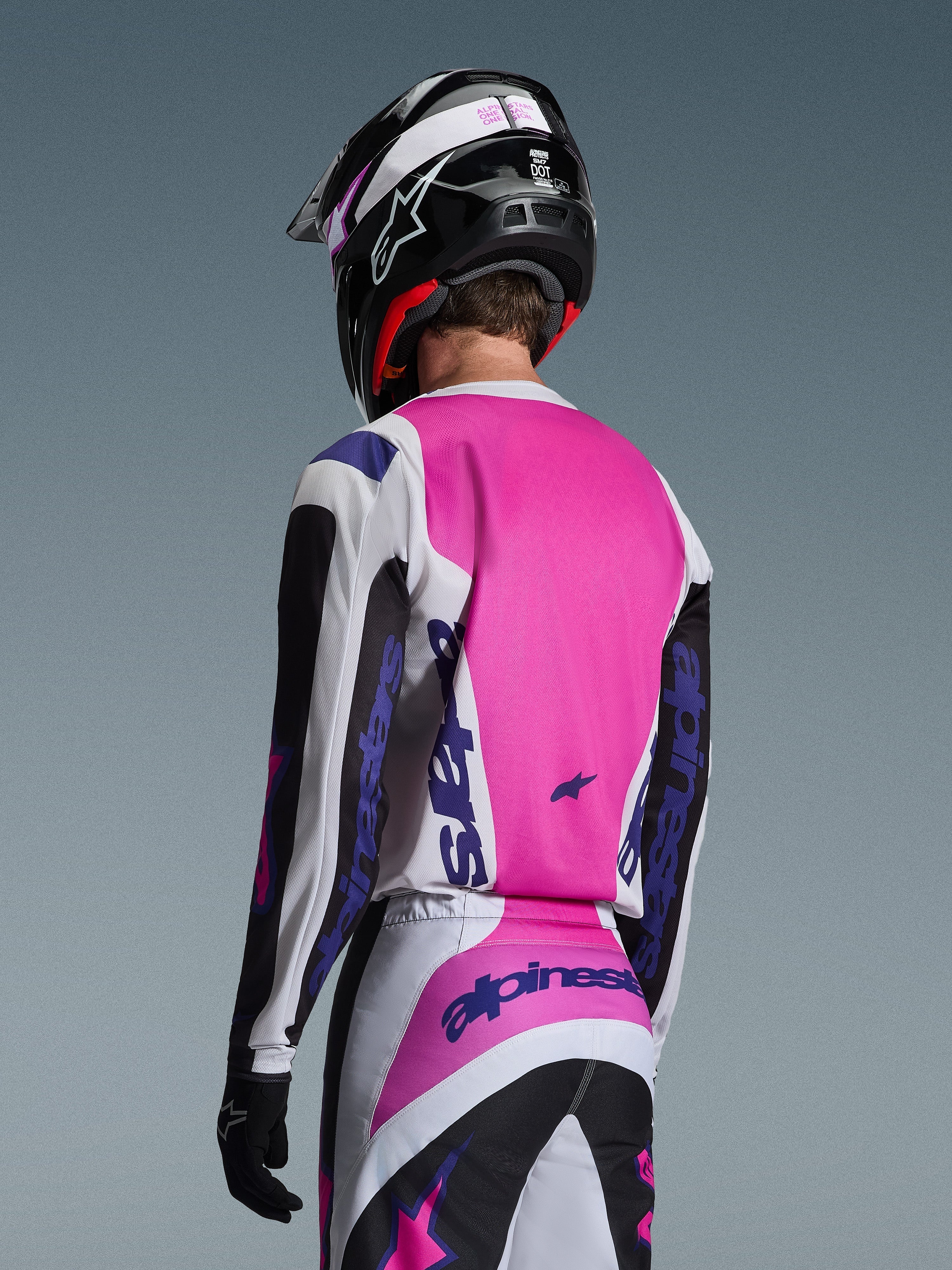 Pilote de motocross vu de dos portant le maillot Alpinestars 2026 Fluid Grid en noir, blanc et violet, pantalon assorti avec marquage audacieux, casque noir avec des touches de rose et gants noirs, tenue coordonnée complète pour la performance tout-terrain.
