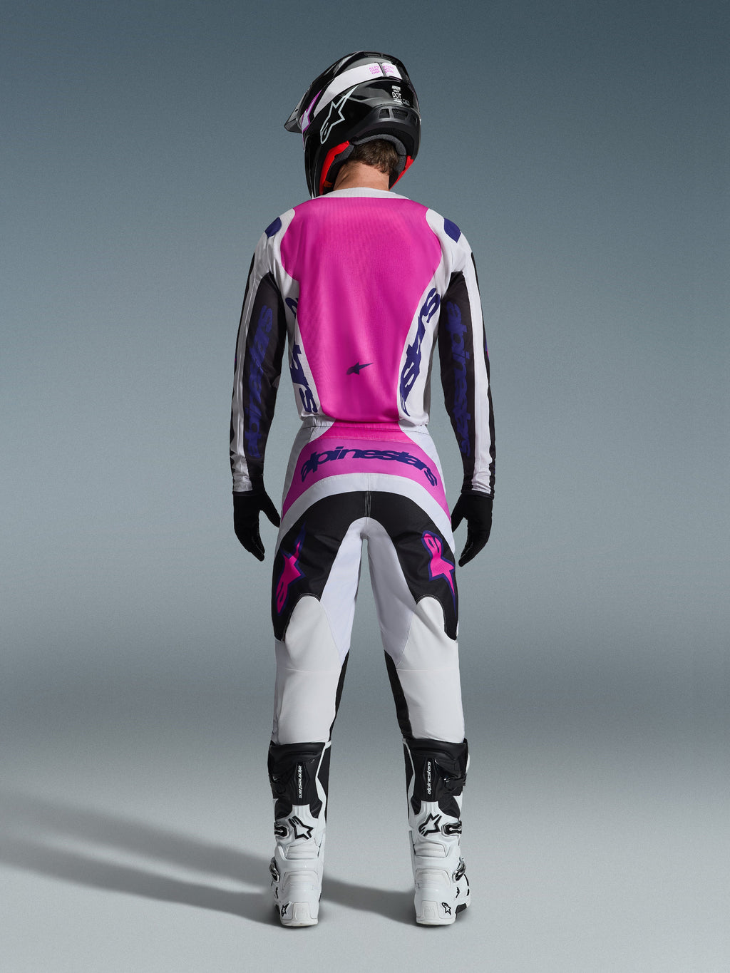 Pilote de motocross vu de dos portant la tenue Alpinestars Fluid Grid 2026, avec un maillot rose vif et blanc aux manches noires, un pantalon blanc assorti avec des panneaux noirs et roses, des logos Alpinestars violets, des bottes de course blanches et noires et un casque coordonné, conçu pour la performance tout-terrain.