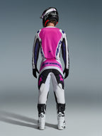 Pilote de motocross vu de dos portant la tenue Alpinestars Fluid Grid 2026, avec un maillot rose vif et blanc aux manches noires, un pantalon blanc assorti avec des panneaux noirs et roses, des logos Alpinestars violets, des bottes de course blanches et noires et un casque coordonné, conçu pour la performance tout-terrain.