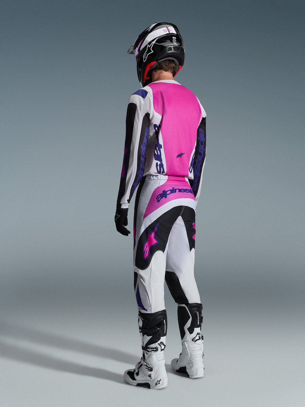 Pilote de motocross vu de dos portant le maillot Alpinestars Fluid Grid 2026 et le pantalon assorti, gris clair et noir avec un empiècement arrière rose vif et des logos violets, complété par un casque noir et des bottes blanches.