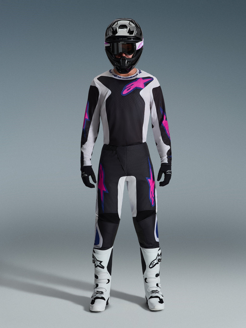 Pilote de motocross portant un équipement de course complet, maillot Alpinestars Fluid Grid 2026 et pantalon assorti en noir et gris clair avec des accents violet vif, casque noir avec lunettes violettes, bottes de motocross blanches et gants noirs, tenue coordonnée pour la performance tout-terrain