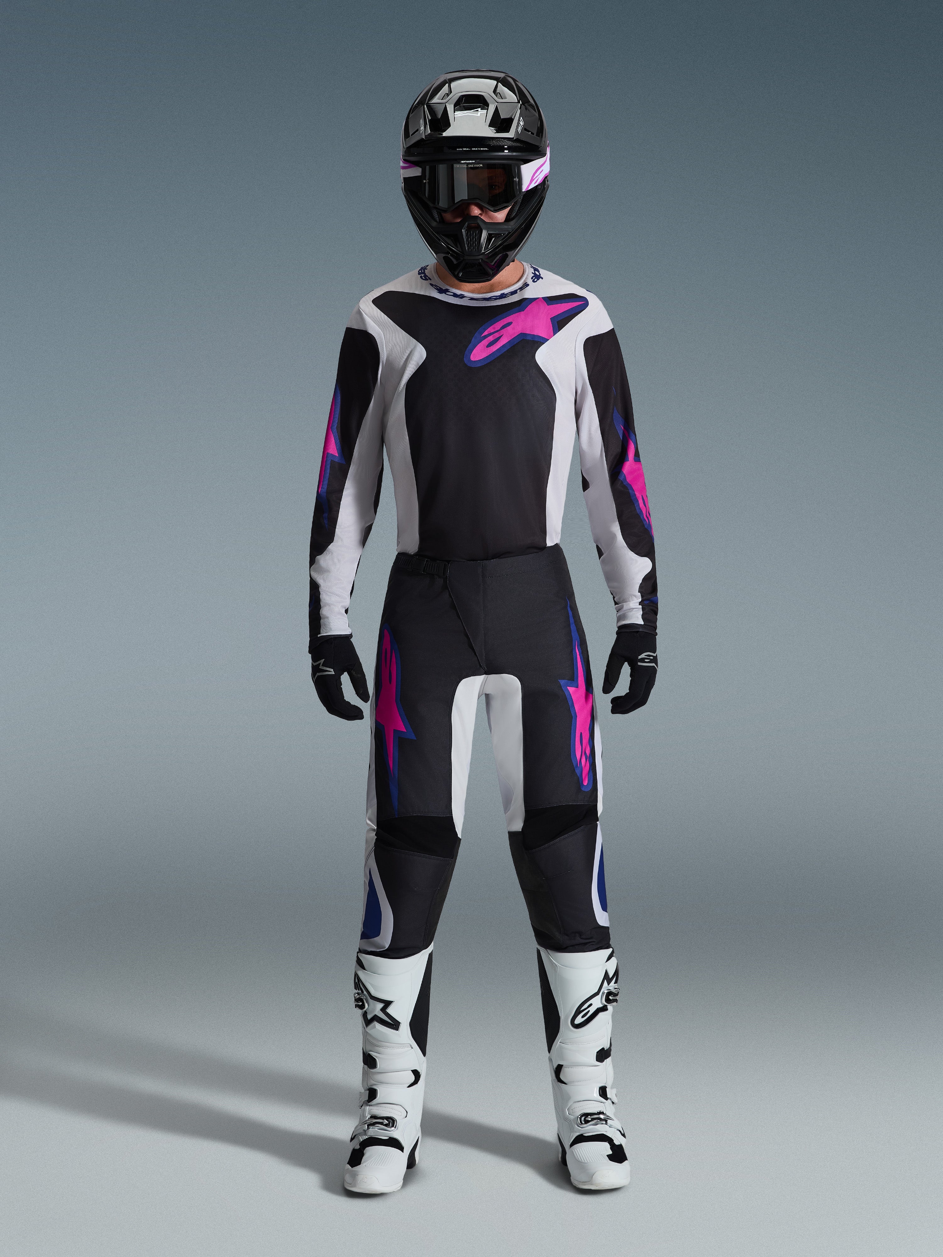 Pilote de motocross portant un équipement de course complet, maillot Alpinestars Fluid Grid 2026 et pantalon assorti en noir et gris clair avec des accents violet vif, casque noir avec lunettes violettes, bottes de motocross blanches et gants noirs, tenue coordonnée pour la performance tout-terrain