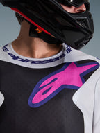 Maillot Alpinestars 2026 Fluid Grid, maillot de motocross, noir, gris clair et violet, gros plan sur la poitrine et le cou avec un grand logo d'étoile rose, col sport logotypé, porté avec un casque noir, équipement de performance tout-terrain.