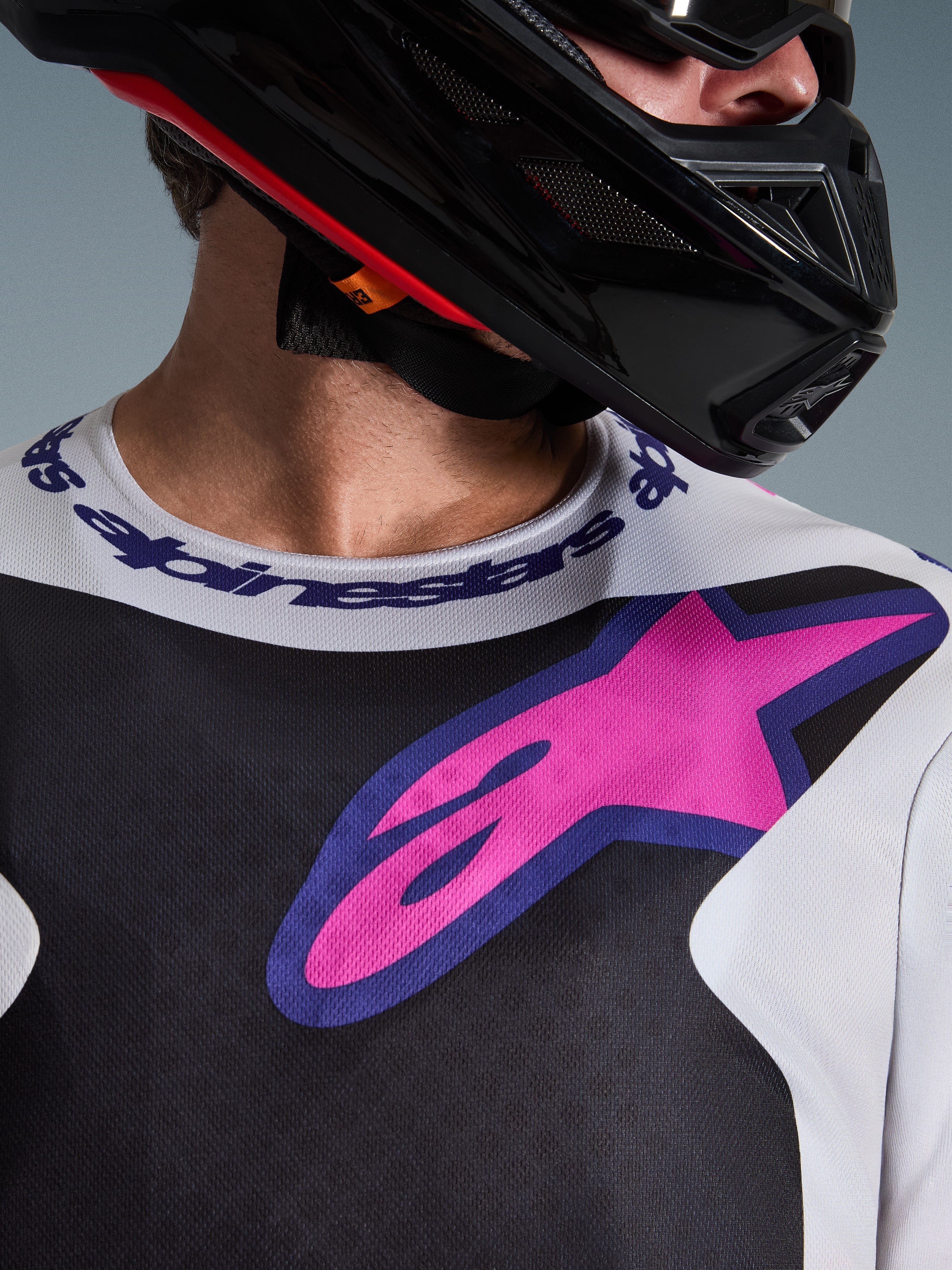 Gros plan sur le Maillot Fluid Grid 2026 d'Alpinestars, maillot de motocross, noir, gris clair et violet, doté d'un logo en forme d'étoile rose-violet vif sur la poitrine et d'un col sport logotypé, porté avec un casque de motocross noir