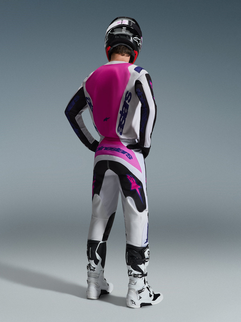 Pilote de motocross de dos portant le maillot Alpinestars Fluid Grid 2026, maillot blanc avec un large panneau rose vif au dos et logos violets, pantalon blanc et noir assorti avec des touches de rose, bottes blanches et casque noir, équipement de course tout-terrain coordonné