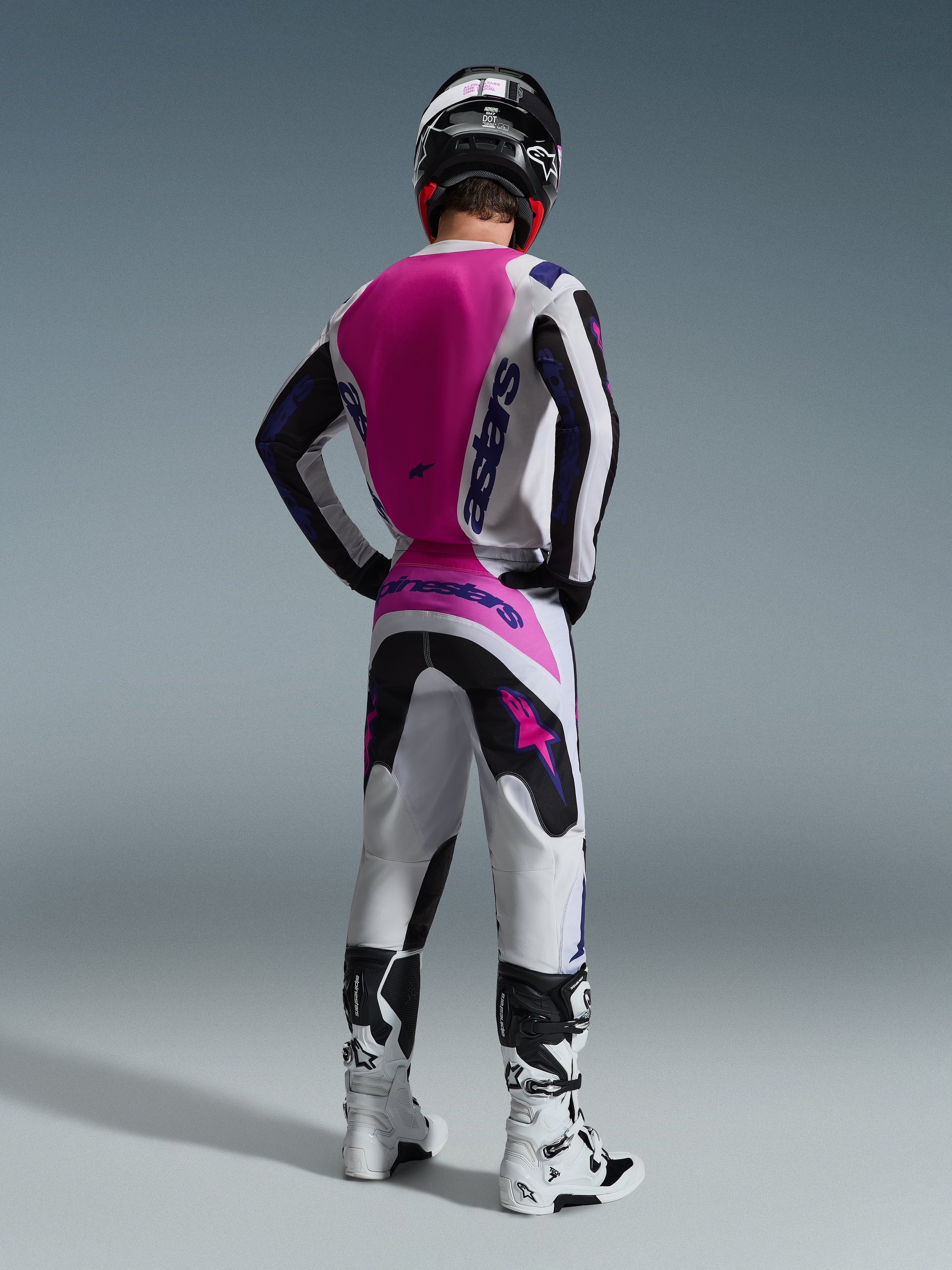 Pilote de motocross de dos portant le maillot Alpinestars Fluid Grid 2026, maillot blanc avec un large panneau rose vif au dos et logos violets, pantalon blanc et noir assorti avec des touches de rose, bottes blanches et casque noir, équipement de course tout-terrain coordonné