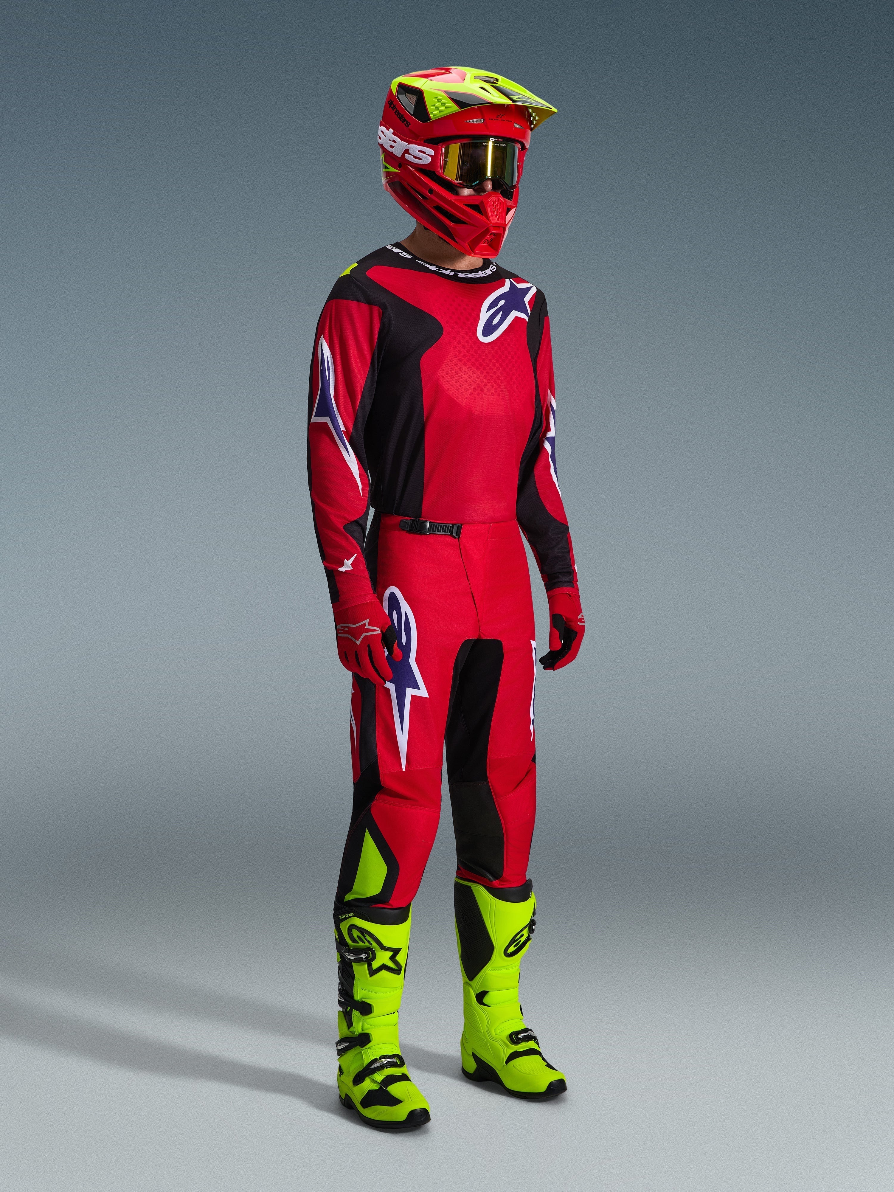 Pilote de motocross portant la tenue complète Alpinestars Fluid Grid 2026 en rouge, noir et violet, comprenant un maillot et un pantalon assortis avec des logos violets, un casque rouge et jaune fluo, et des bottes jaune fluo pour un équipement de course tout-terrain complet.