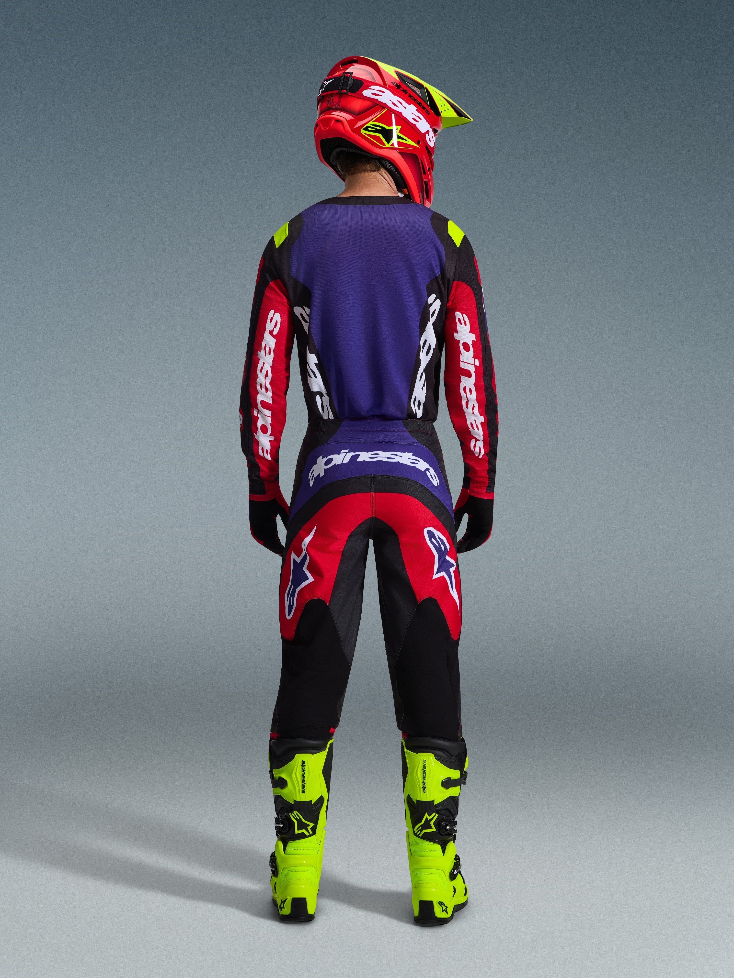 Pilote de motocross vu de l'arrière portant l'ensemble d'équipement Alpinestars Fluid Grid 2026 en coloris Rouge Noir Violet, avec un maillot à panneau arrière violet et manches rouges, un pantalon rouge et noir assorti, des bottes de motocross jaune néon éclatant et un casque rouge et jaune néon.
