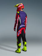 Pilote de motocross portant l'ensemble Alpinestars Fluid Grid 2026, vue de dos, maillot rouge, noir et violet avec logos blancs, pantalon rouge et noir avec accents violets, coordonné avec un casque rouge et jaune fluo et des bottes jaune fluo, équipement de course tout-terrain complet
