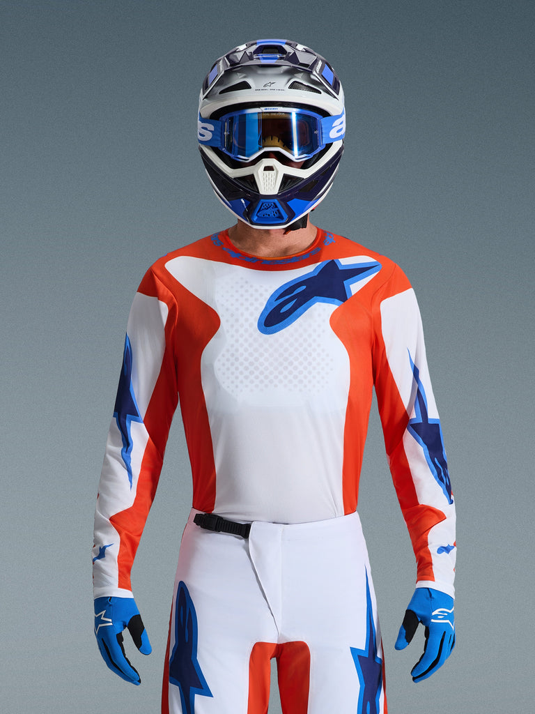 Pilote de motocross portant le maillot Fluid Grid 2026 d'Alpinestars en Orange Ucla Blue, avec torse blanc, panneaux latéraux orange et logos bleus, coordonné avec un pantalon blanc et bleu, des gants bleus et un casque blanc et bleu pour un look de course professionnel.