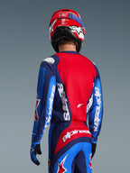 Vue arrière d'un pilote de motocross portant le maillot Alpinestars Fluid Grid 2026 en bleu et rouge, avec des logos blancs sur les manches et le dos, associé à un pantalon de motocross assorti et un casque rouge pour une tenue de course tout-terrain complète