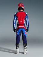 Vue arrière d'un pilote de motocross portant le maillot Alpinestars Fluid Grid 2026 bleu et rouge, doté d'un large panneau dorsal rouge et de manches bleues avec marquage Alpinestars blanc, associé à un pantalon assorti, des bottes blanches et noires et un casque rouge pour une tenue de course tout-terrain complète.