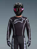 Maillot Alpinestars Fluid Apex 2026, maillot de motocross, noir et gris, avec un grand logo Astars blanc sur la poitrine et des détails graphiques gris sur les manches, porté par un pilote avec des lunettes roses et un casque noir, équipement de course tout-terrain coordonné