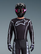 Maillot Alpinestars Fluid Apex 2026, maillot de motocross, noir et gris, avec un grand logo Astars blanc sur la poitrine et des détails graphiques gris sur les manches, porté par un pilote avec des lunettes roses et un casque noir, équipement de course tout-terrain coordonné