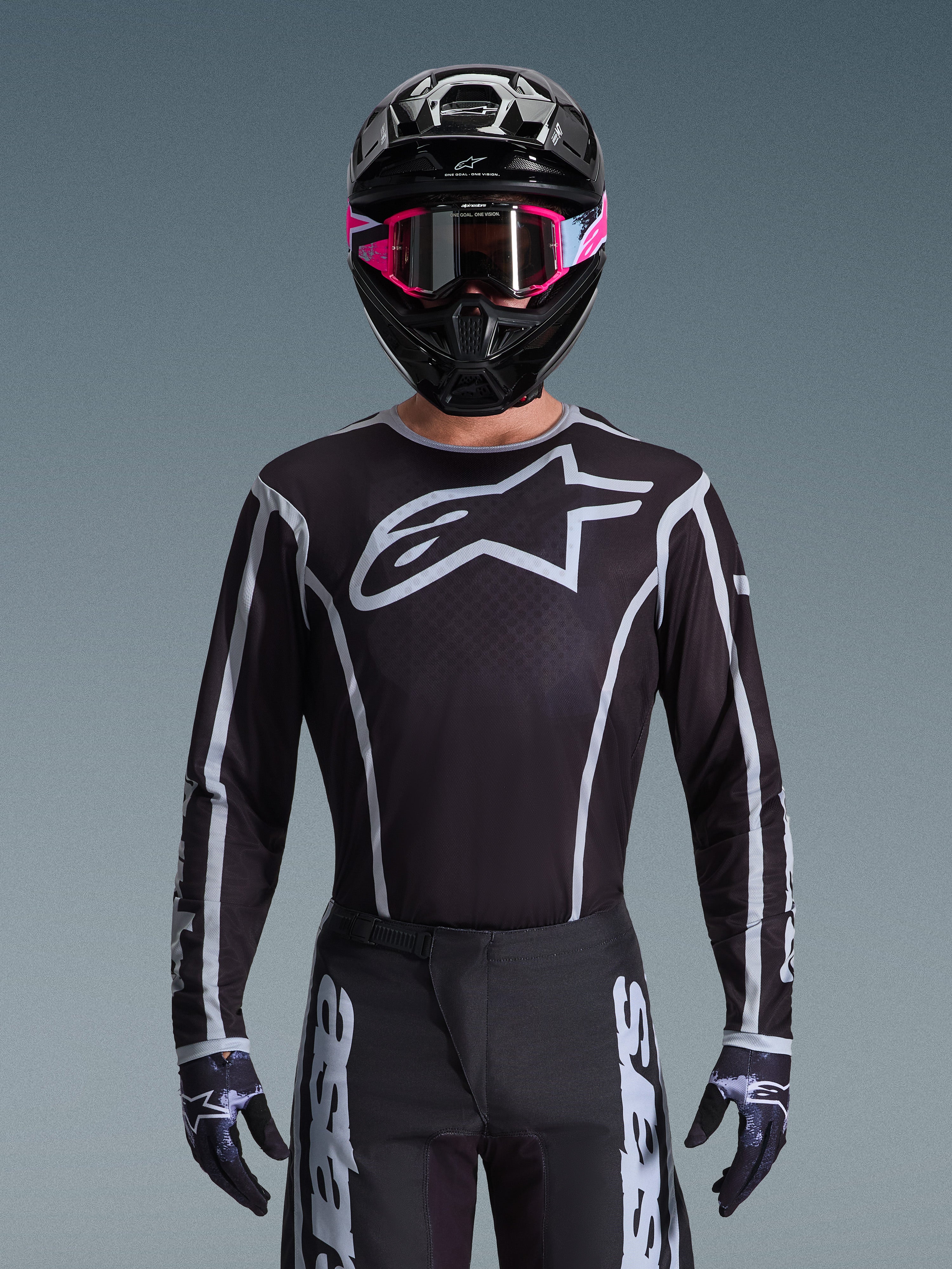 Maillot Alpinestars Fluid Apex 2026, maillot de motocross, noir et gris, avec un grand logo Astars blanc sur la poitrine et des détails graphiques gris sur les manches, porté par un pilote avec des lunettes roses et un casque noir, équipement de course tout-terrain coordonné