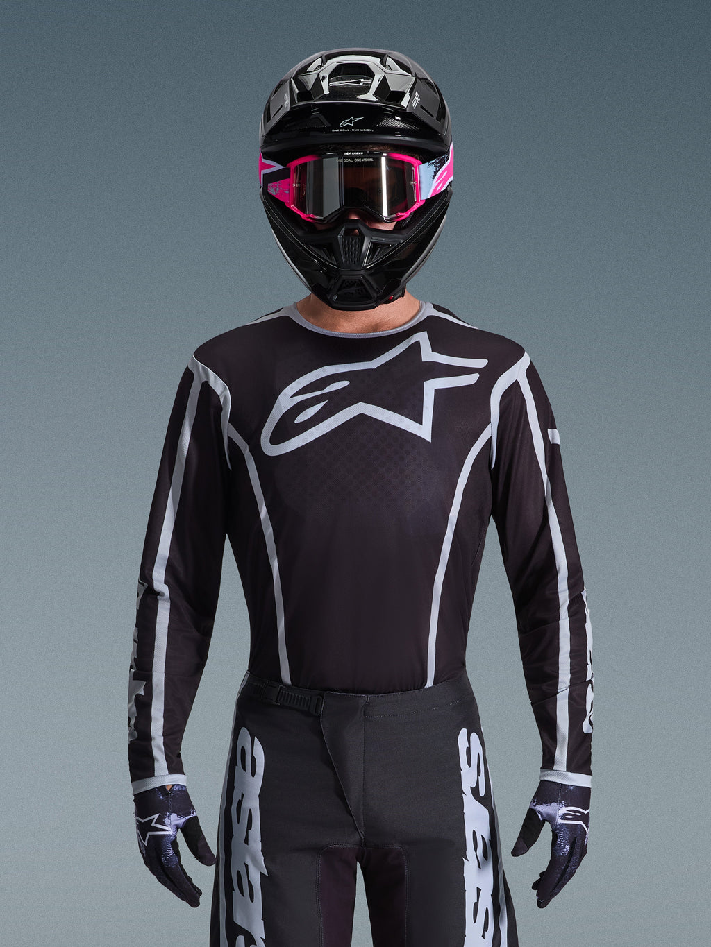 Ensemble d'équipement Alpinestars Fluid Apex 2026, maillot et pantalon de motocross noir et blanc, avec un grand logo sur la poitrine et des lignes géométriques, porté avec un casque noir et des lunettes roses, équipement de performance pour le motocross tout-terrain