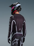 Pilote de motocross de dos portant le maillot Alpinestars Fluid Apex 2026 noir et gris, pantalon de motocross assorti avec accents blancs, casque noir avec logo rose, tenue coordonnée pour la course tout-terrain