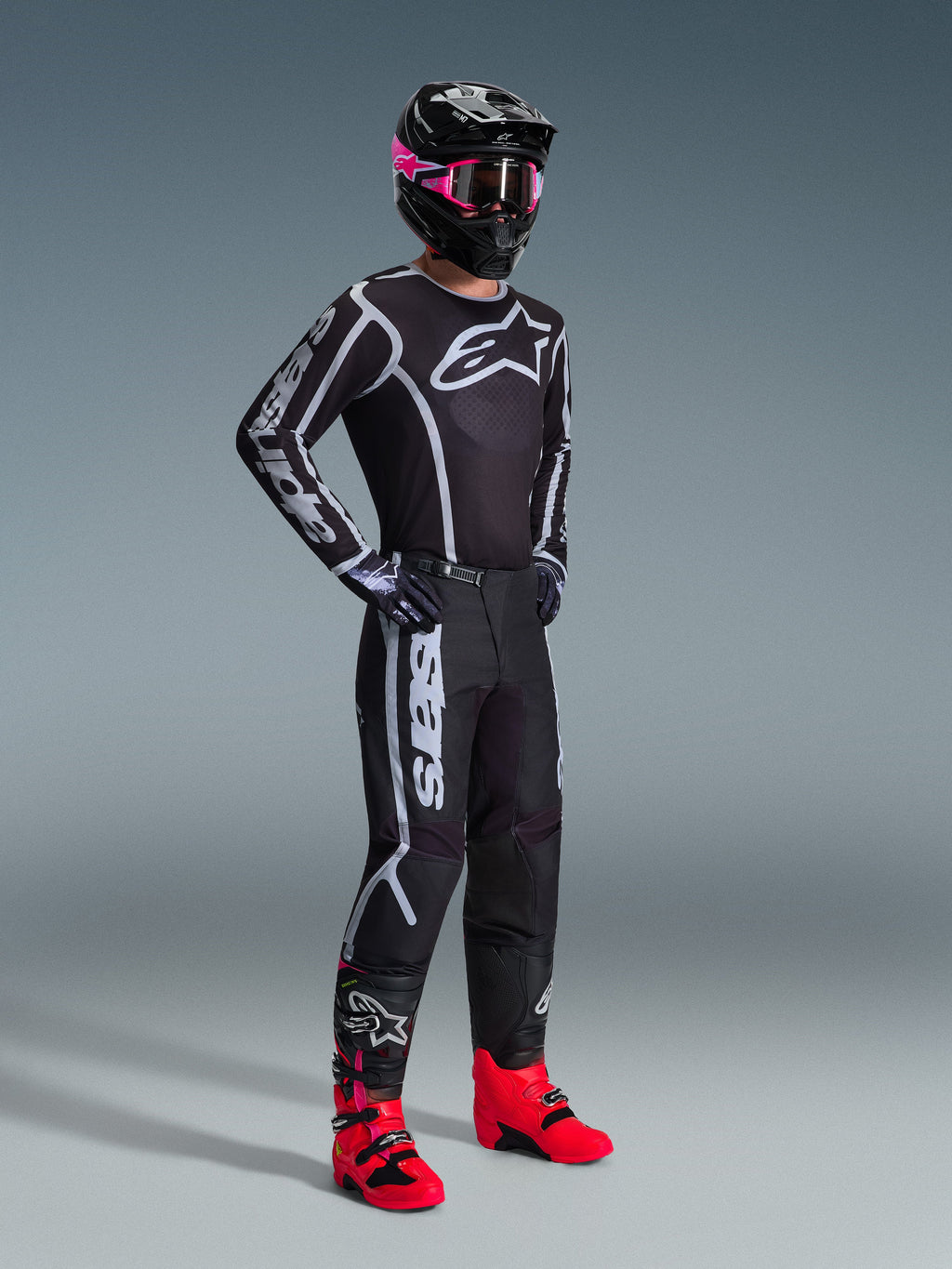 Pilote de motocross portant le maillot 2026 Fluid Apex, noir et gris, associé à un pantalon assorti, un casque noir avec des lunettes roses et des bottes rouge vif, avec le logo Alpinestars blanc sur la poitrine et les manches, tenue de course tout-terrain professionnelle coordonnée.