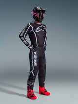 Motocross rider wearing 2026 Fluid Apex Jersey, Black and Gray, paired with matching pants, black helmet with pink goggles, and bright red boots, featuring bold white Alpinestars branding on the chest and sleeves, coordinated professional off-road racing outfit.