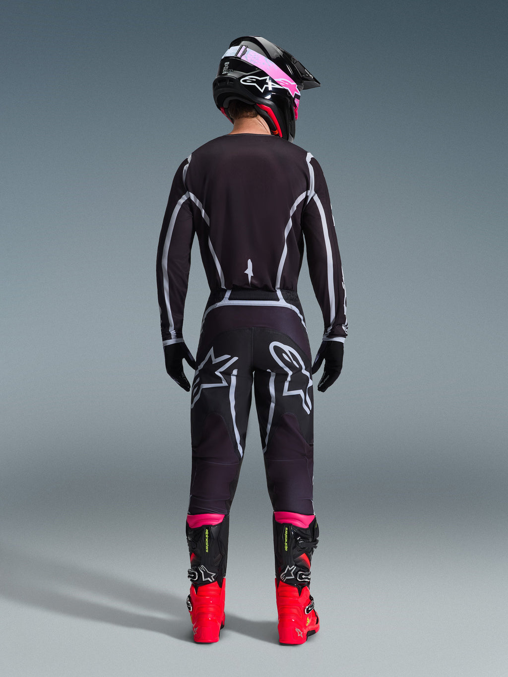 Pilote de motocross vu de dos portant l'ensemble d'équipement Alpinestars 2026 Fluid Apex, maillot et pantalon noirs avec lignes géométriques grises et logos étoilés, casque noir avec accents roses, bottes rouge vif et noires, tenue de motocross coordonnée