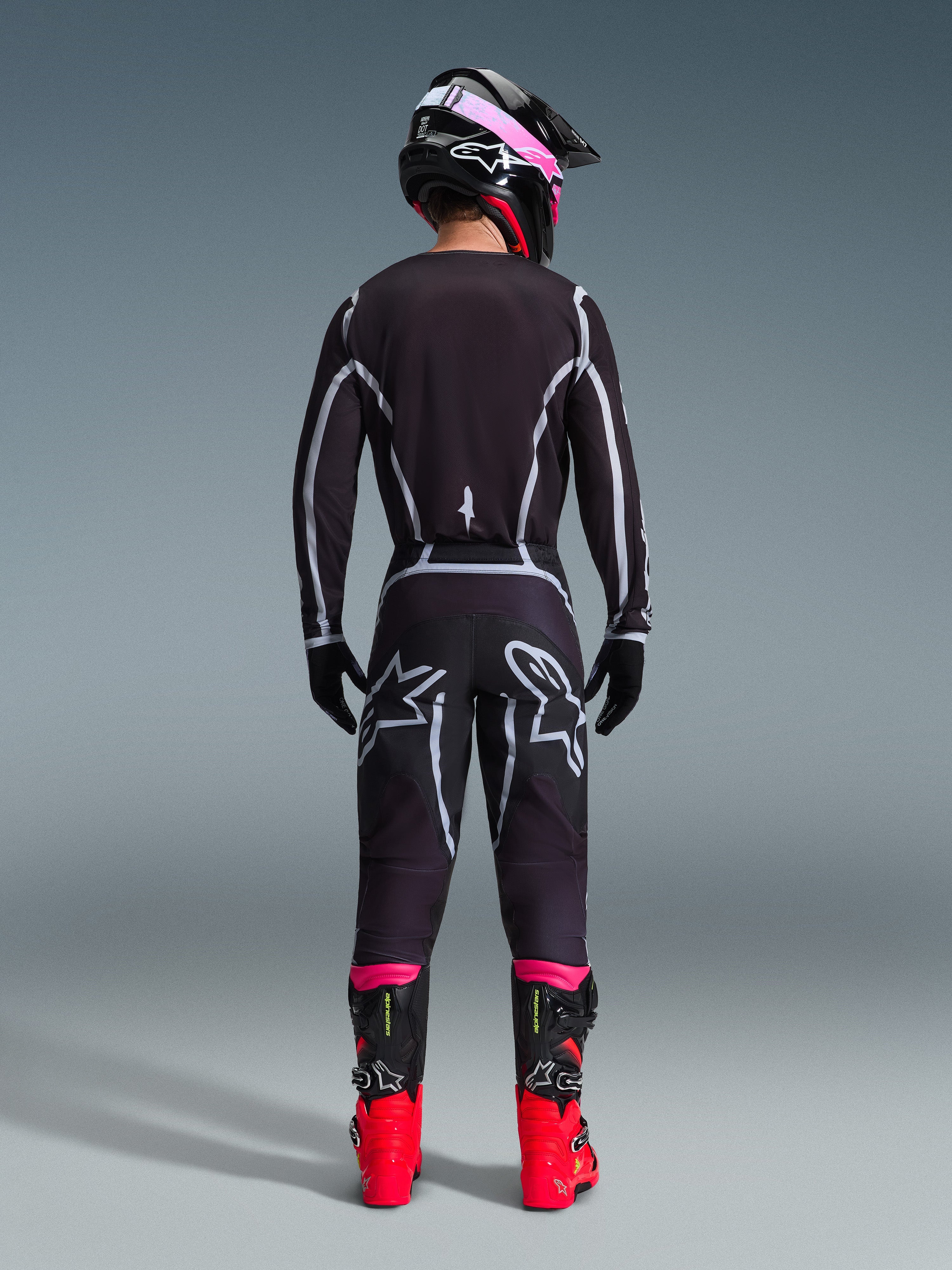 Pilote de motocross vu de dos portant l'ensemble d'équipement Alpinestars 2026 Fluid Apex, maillot et pantalon noirs avec lignes géométriques grises et logos étoilés, casque noir avec accents roses, bottes rouge vif et noires, tenue de motocross coordonnée