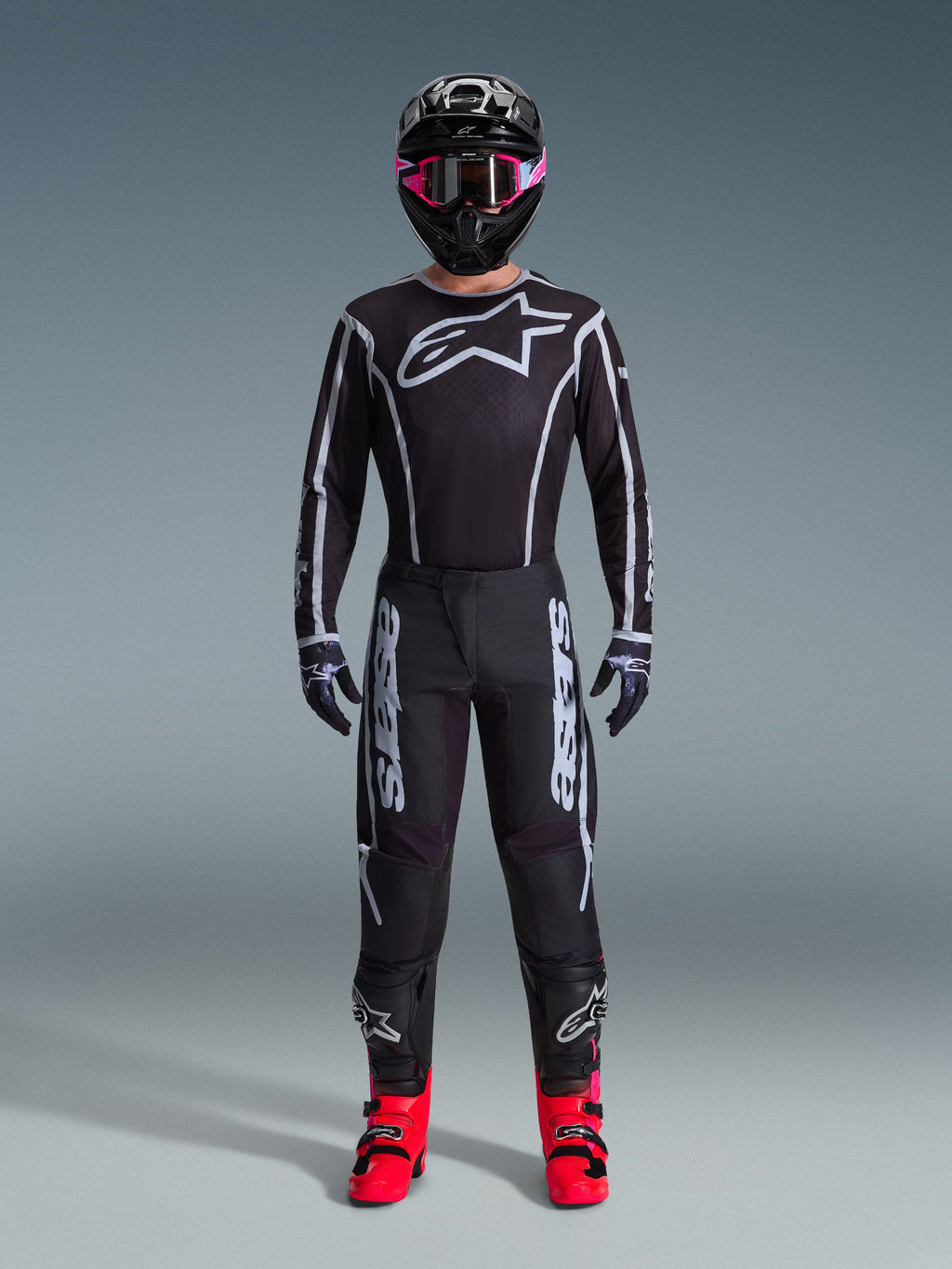 Pilote de motocross portant le maillot Alpinestars Fluid Apex 2026, noir et gris, grand logo Astars blanc sur la poitrine, lignes géométriques blanches, pantalon noir assorti, casque noir avec lunettes roses et bottes rose vif, tenue de course de motocross complète