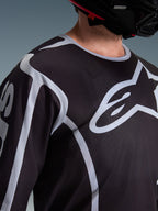 Maillot Alpinestars Fluid Apex 2026, maillot de motocross, noir et gris, avec un grand logo Alpinestars sur la poitrine, col sport, porté avec un casque noir, équipement de performance pour la conduite tout-terrain