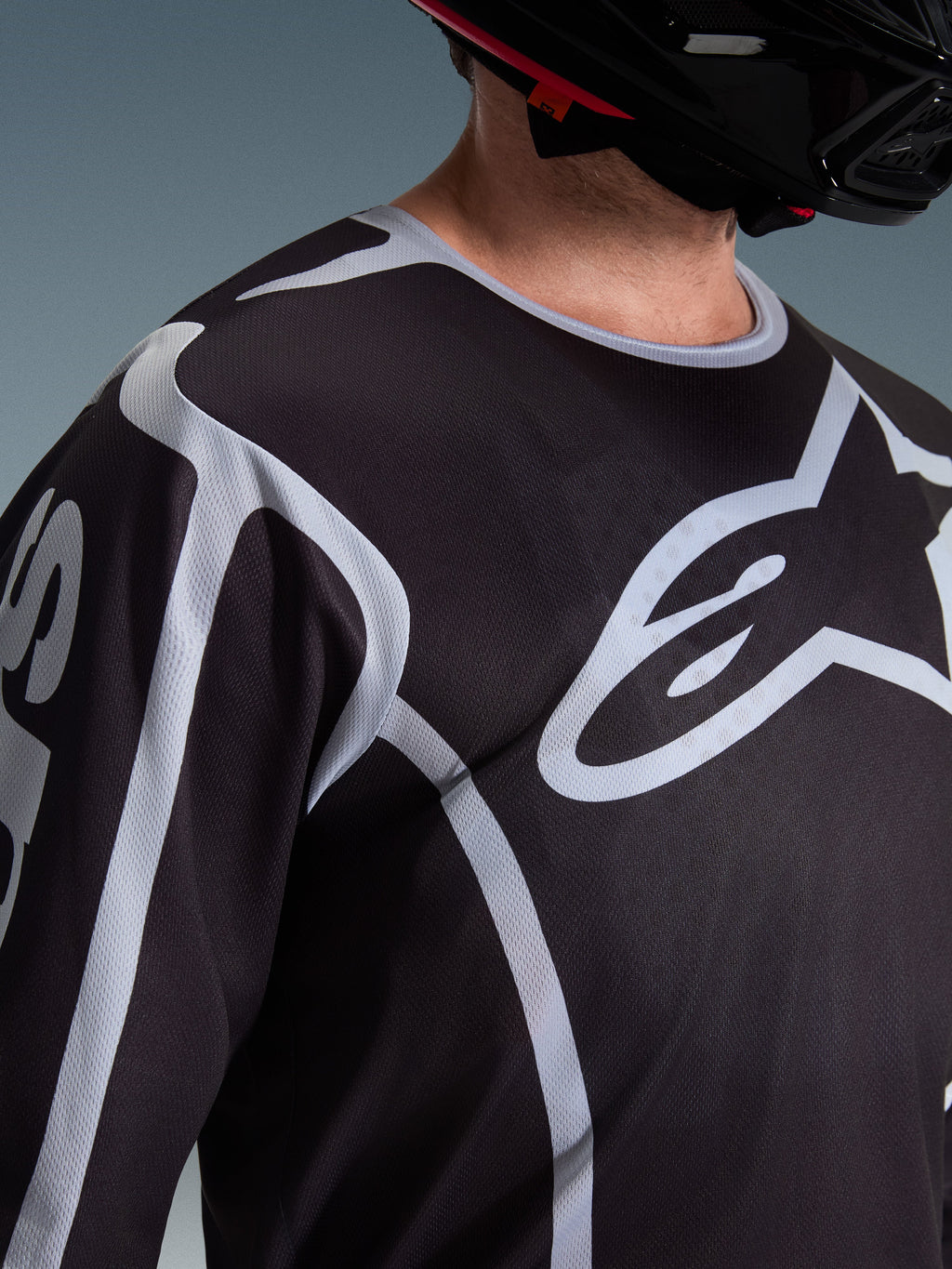 Gros plan d'un pilote portant le maillot Alpinestars 2026 Fluid Apex noir et gris, mettant en avant le grand logo Astars blanc sur la poitrine et la construction technique en mesh, avec un casque de motocross noir.