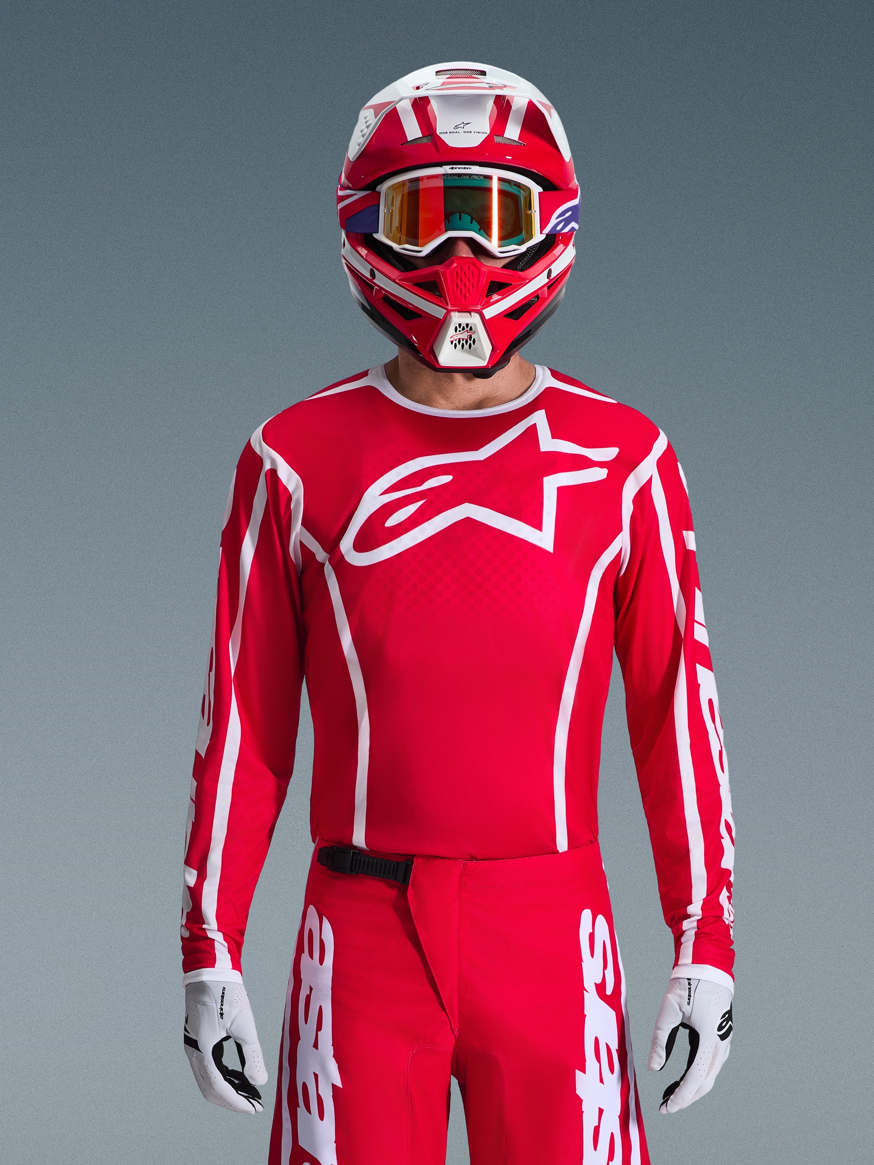 Motocross rider wearing Alpinestars 2026 Fluid Apex Gear Set, Red jersey with large white star logo and white piping, matching red pants with white accents, red and white helmet with purple highlights and orange-tinted goggles, white and black gloves, coordinated outfit for off-road racing