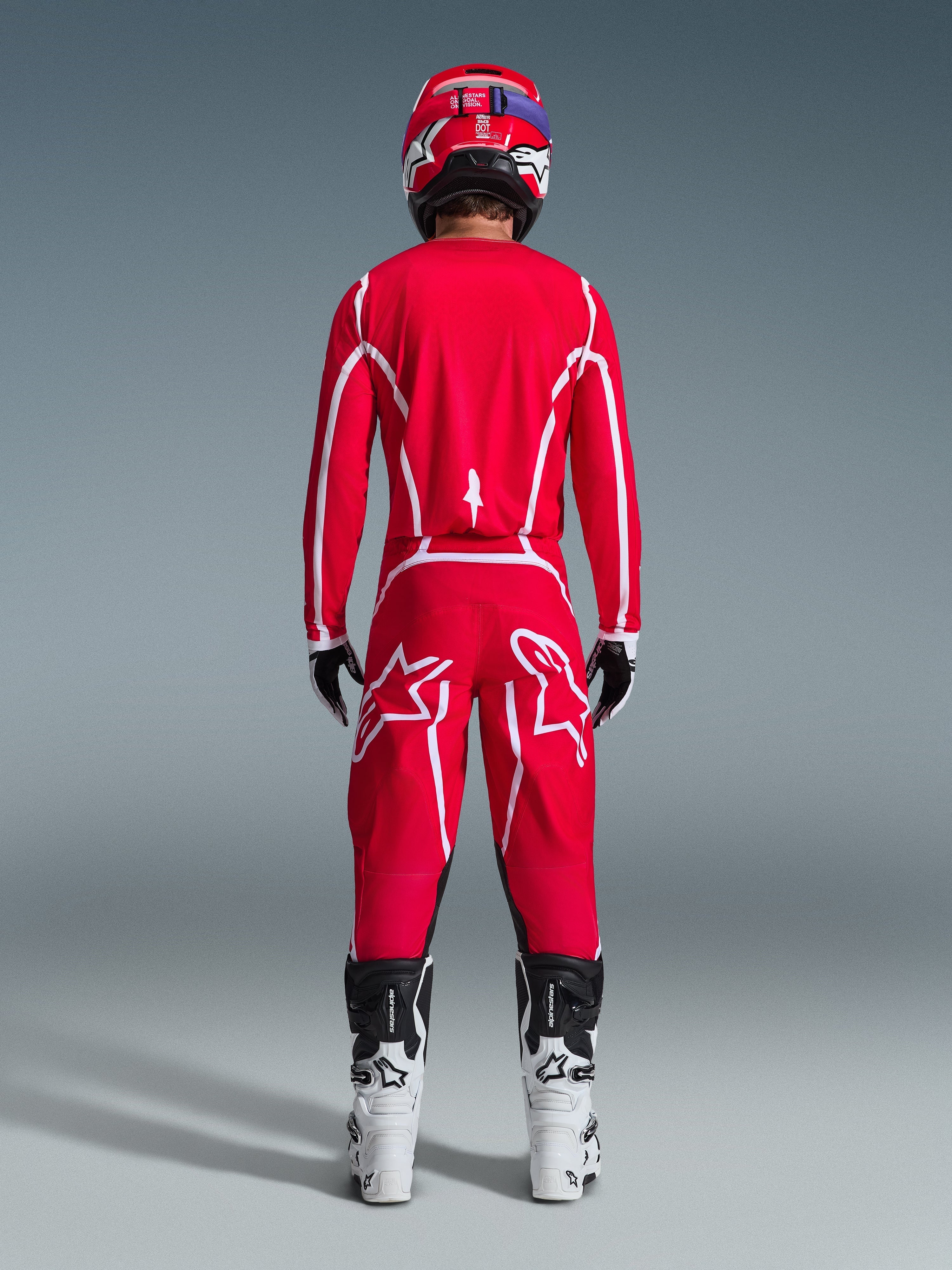 Motocross rider viewed from behind wearing the Alpinestars 2026 Fluid Apex Jersey in Red and White, matching red pants with prominent white star logos, a red helmet, white and black motocross boots, and black and white gloves, complete kit for off-road racing