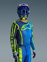 Pilote de motocross portant le maillot Alpinestars 2026 Fluid Apex en bleu et jaune fluo, pantalon assorti bleu et jaune fluo, casque violet et jaune avec lunettes bleues et gants coordonnés, équipement complet de course tout-terrain.