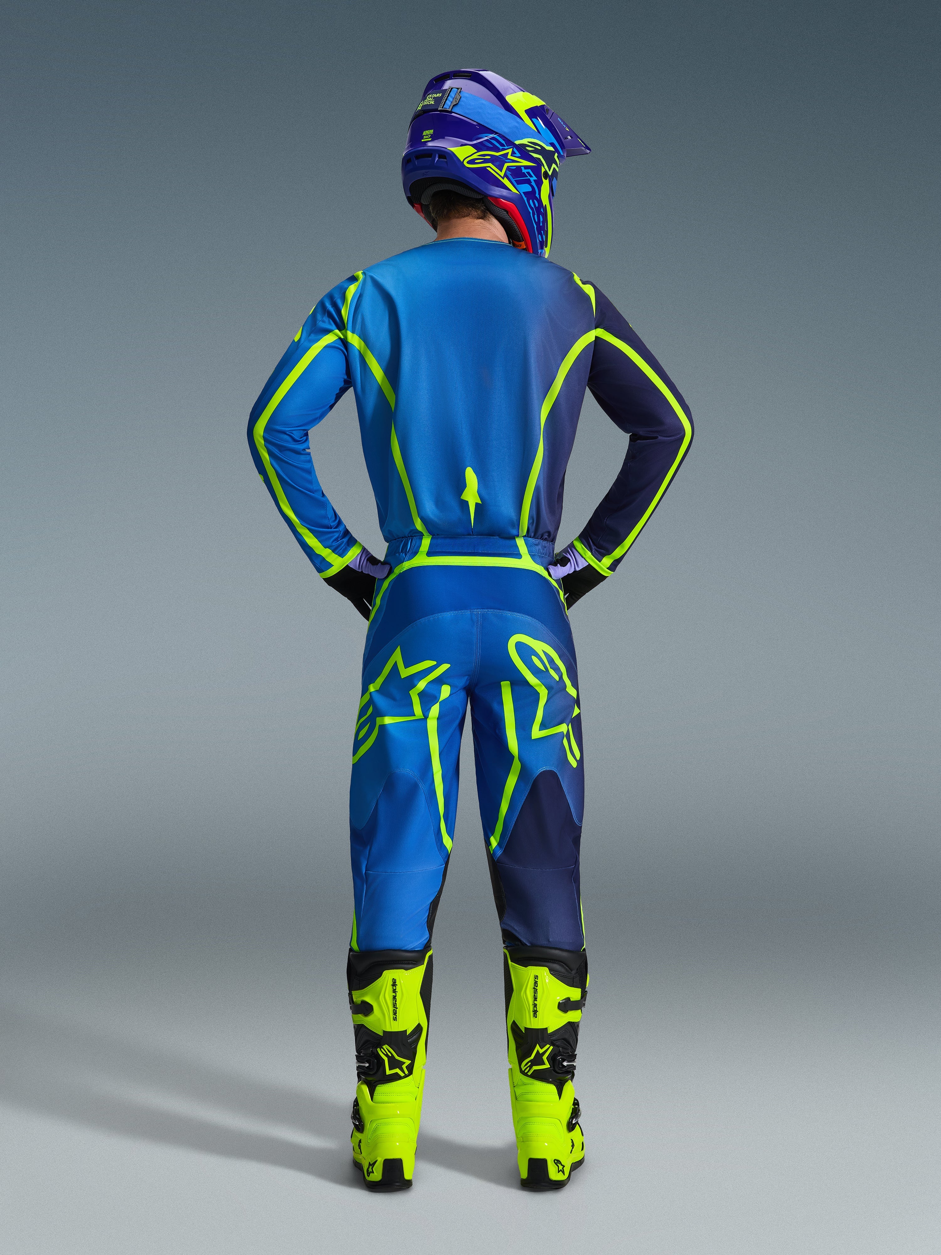 Alpinestars 2026 Fluid Apex Gear Set, back view of a motocross rider wearing a blue and navy jersey and matching pants with vibrant neon green accents, neon yellow boots, and a purple helmet, professional off-road racing outfit.