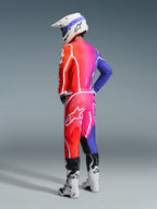 Motocross rider wearing the Alpinestars 2026 Fluid Apex Gear Set, vibrant gradient transition from orange to pink and purple with bold white branding and star logos, paired with a white helmet, blue goggles, and white boots, seen from a rear three-quarter angle in a studio setting.