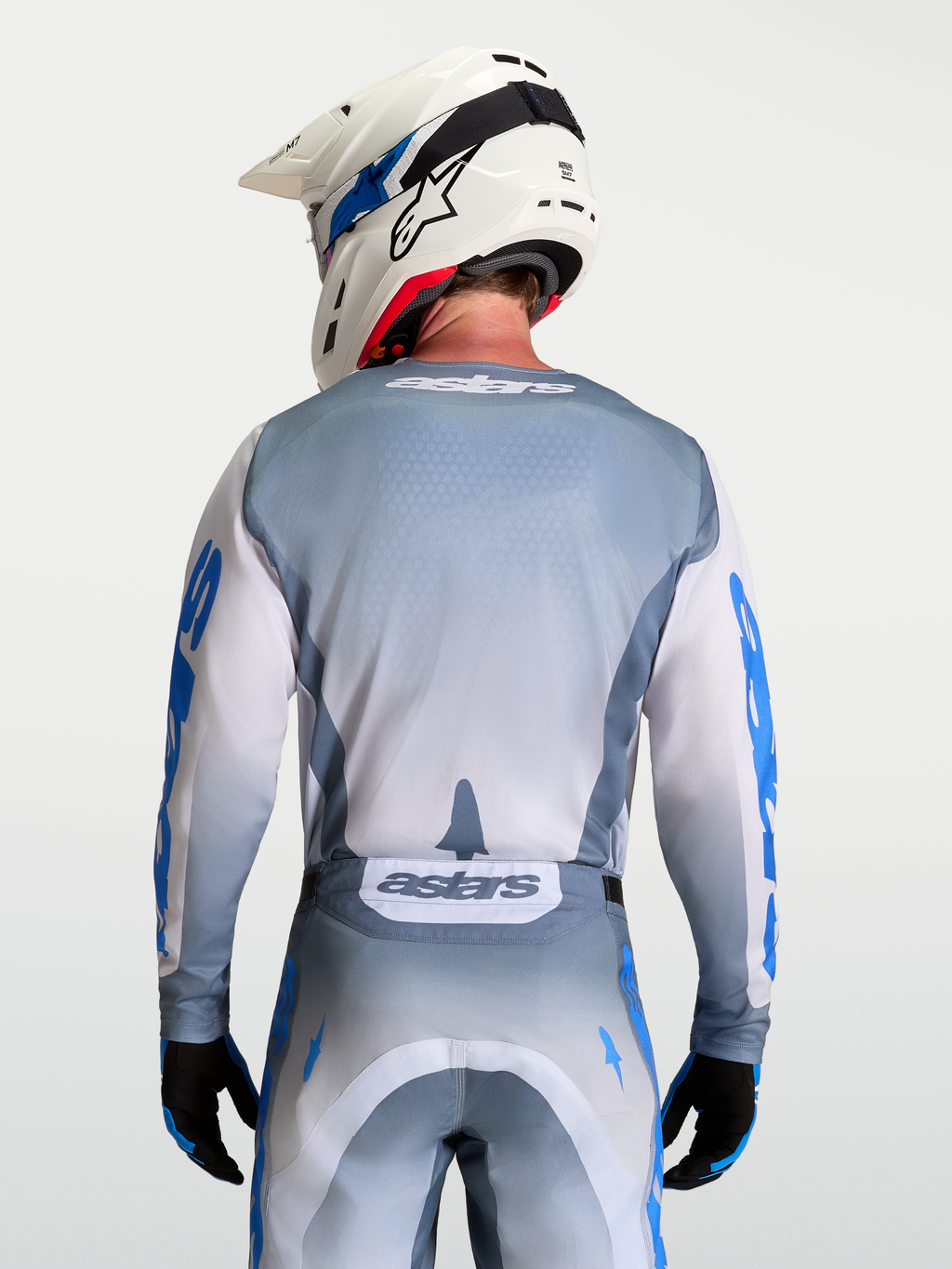 Alpinestars Maillot Racer Amber, maillot de motocross gris et blanc, vue de dos sur mannequin avec casque, graphismes bleu UCLA et lilas, coupe ergonomique racing, tissu poly-matière évacuant l'humidité