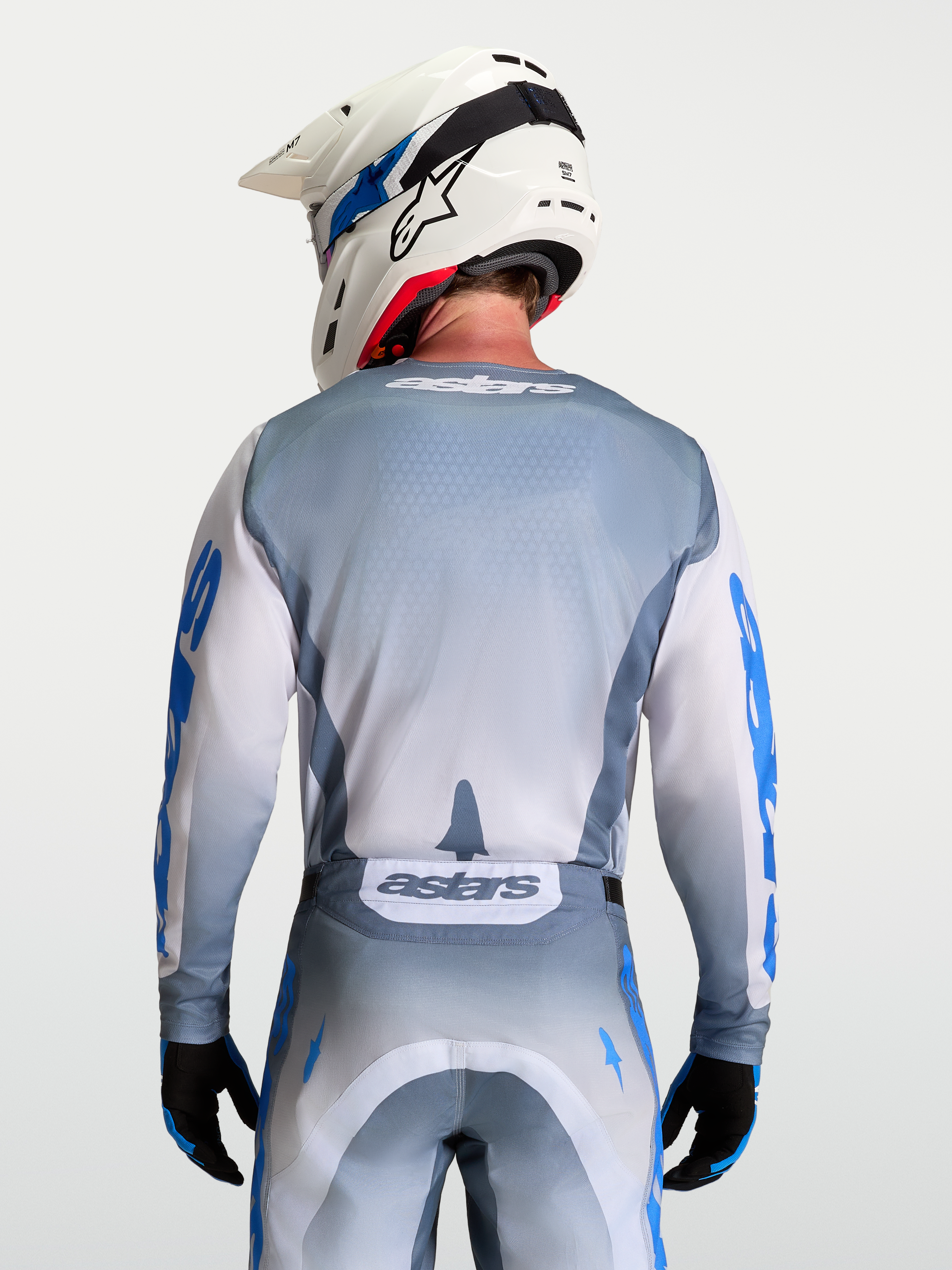 Alpinestars Maillot Racer Amber, maillot de motocross gris et blanc, vue de dos sur mannequin avec casque, graphismes bleu UCLA et lilas, coupe ergonomique racing, tissu poly-matière évacuant l'humidité