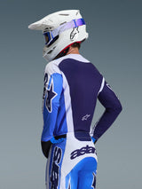 Pilote de motocross portant le maillot Alpinestars 2026 Racer Air Portl en bleu UCLA et blanc, vue arrière mettant en valeur le panneau dorsal en maille respirante bleu marine, porté avec un pantalon assorti et un casque Alpinestars blanc avec des lunettes bleues, tenue de motocross coordonnée.