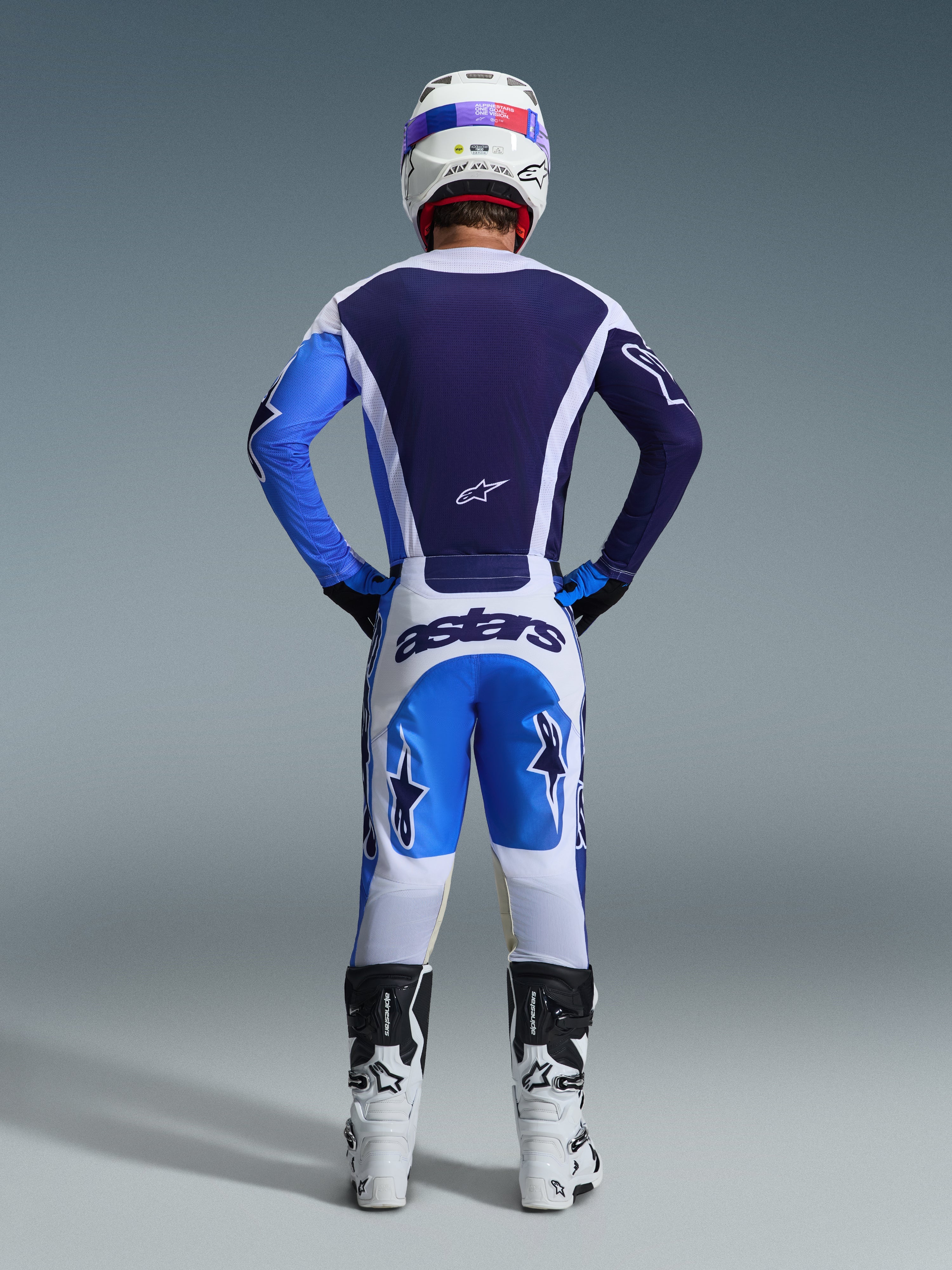 Motocross rider seen from behind wearing the Alpinestars 2026 Racer Air Portl Gear Set, Navy and bright blue jersey with white accents, white pants with blue panels and navy 'astars' branding, white and black motocross boots, white racing helmet, coordinated outfit for off-road performance
