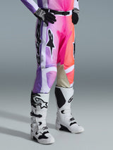 Pantalon Alpinestars Racer Air Portl 2026, pantalon de motocross, graphisme multicolore avec dégradés de violet, rose et orange, porté avec des bottes de motocross blanches et noires et des gants noirs, doté d'une coupe de course et de renforts intérieurs aux genoux en cuir beige.